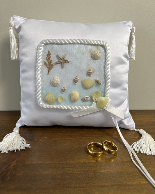 Almofadinha para alianças em cetim com conchas, areia e franjas nas pontas. Modelo Praia da Etiquette, ideal para casamentos à beira-mar.