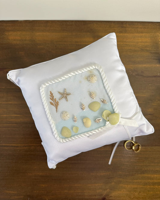 Almofadinha para alianças em cetim com conchas, areia e franjas nas pontas. Modelo Praia da Etiquette, ideal para casamentos à beira-mar.